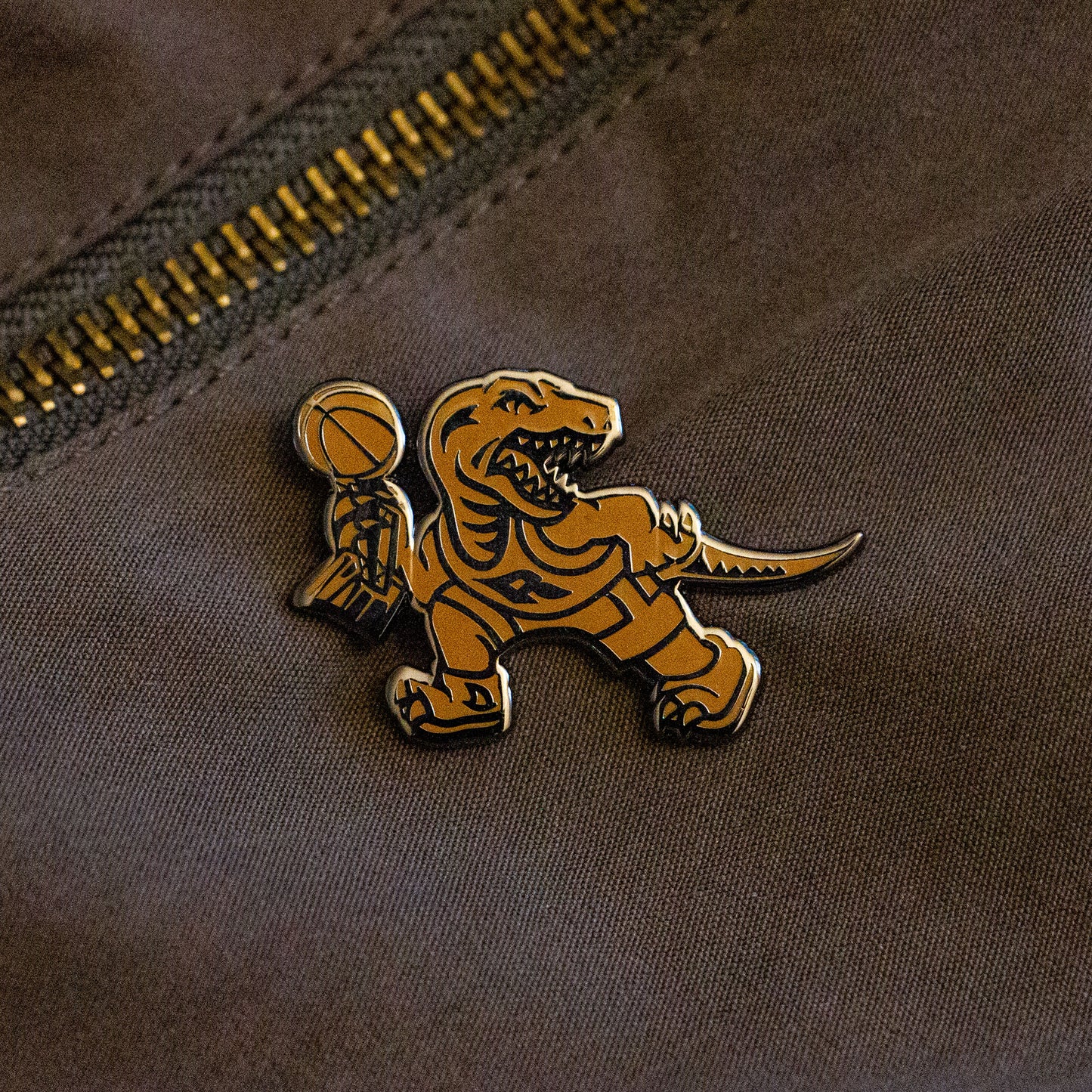 A gold hard enamel pin of the Toronto Raptors mascot logo holding the championship trophy. This design is to commemorate the NBA Championship won by the Raptors in the 2018-2019 season.