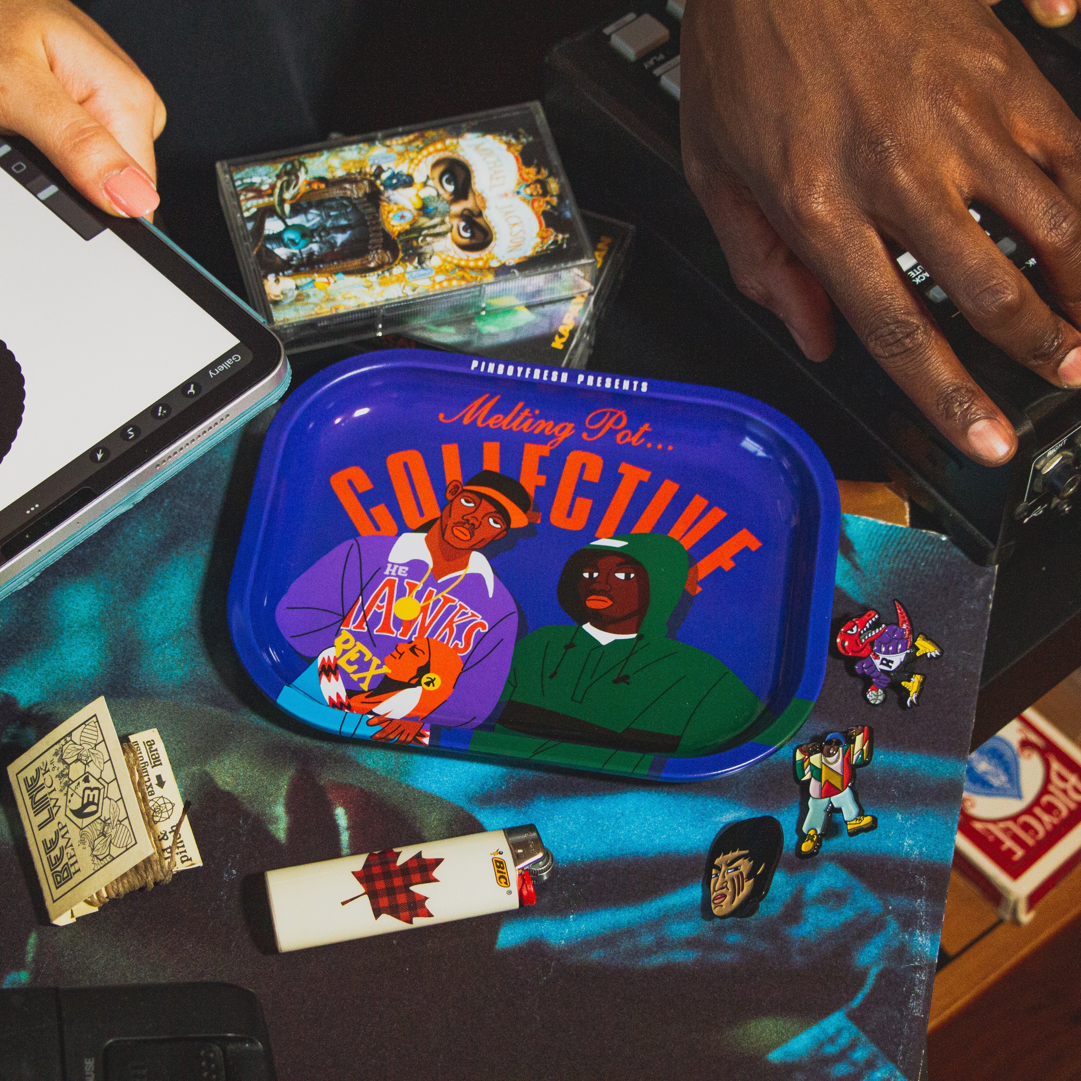 A custom Mobb Deep rolling tray by Pin Boy Fresh is on a table on top of vinyl records with various items around it, from enamel pins, cassettes, lighter, matches, and a drum machine.