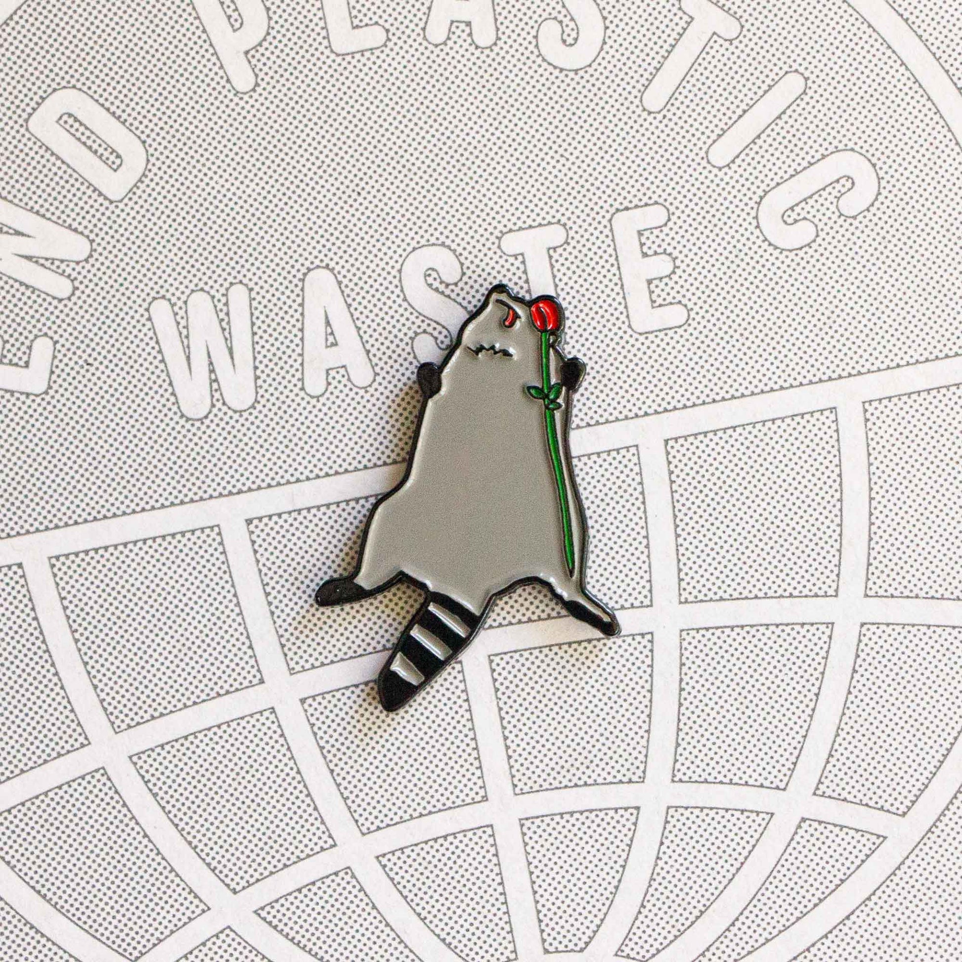 An enamel pin that is a parody of a Toronto raccoon roadkill that went viral from people throwing a memorial for it. The raccoon is laying lifeless with it's tongue out, holding a rose.